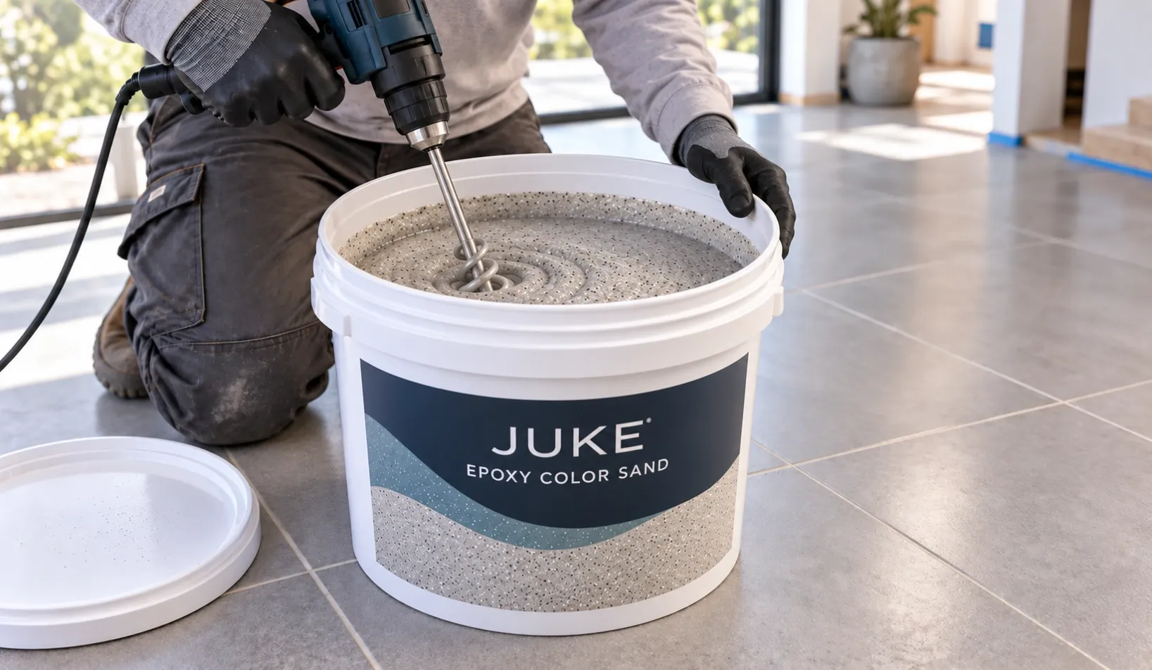 Installer kneeling on a tiled floor and mixing epoxy color sand in a white JUKE bucket with an electric paddle mixer.