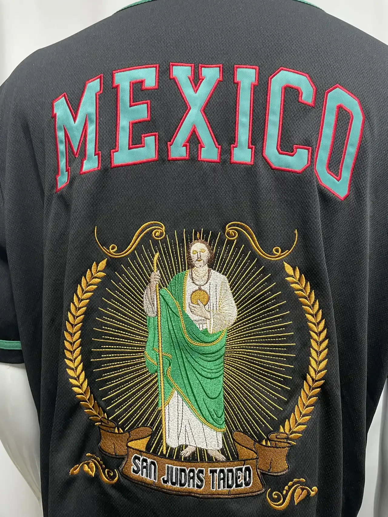 Back of black baseball jersey with raised San Judas figure embroidery inside a golden wheat wreath