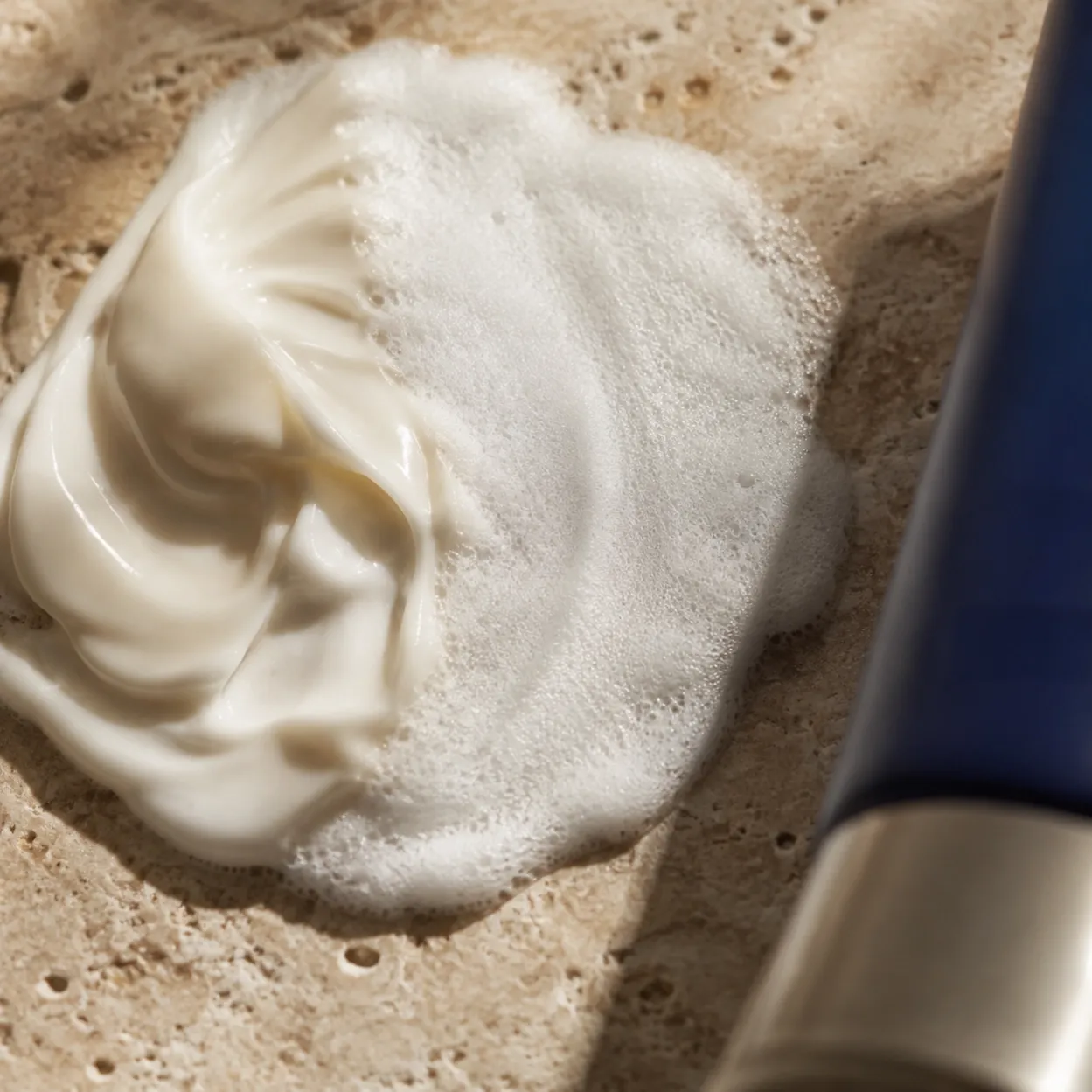 Glossy ivory cleansing cream opening into fine dense microfoam on travertine