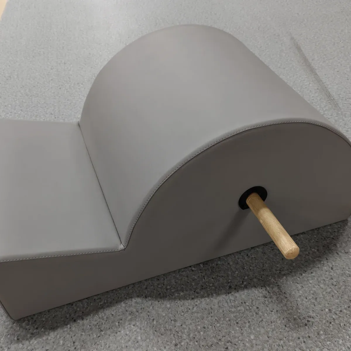 Front three-quarter view of the grey upholstered Pilates spine corrector, showing the gradual arc, the flat pad and a wooden bar projecting at the side