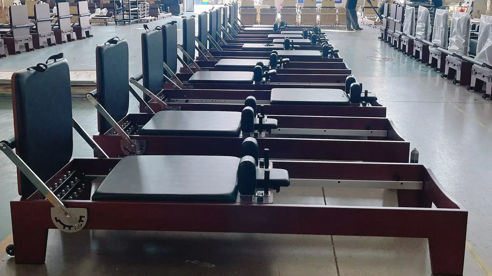 A long row of Pilates reformers with walnut-tone frames, black padding and metal footbars on a workshop floor