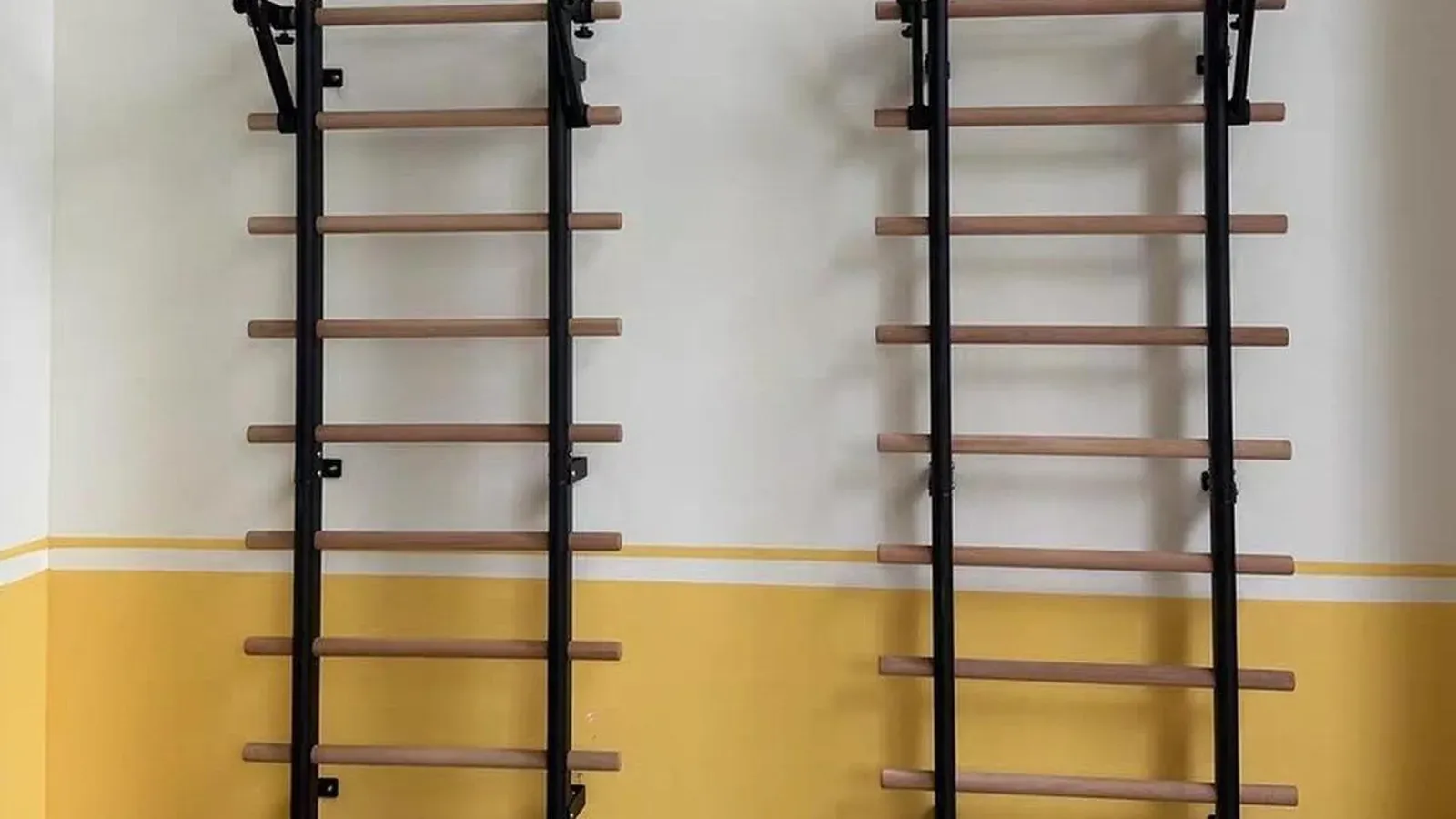 Two Swedish ladder stall bars with black metal uprights and light wooden rungs mounted on a wall, each with a removable pull-up bar across the top