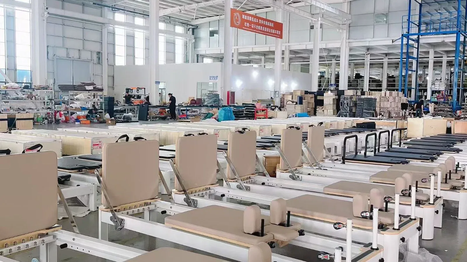 Rows of white-framed Pilates reformers with light coffee leather upholstery on their carriages, in a workshop with wooden shipping crates behind them