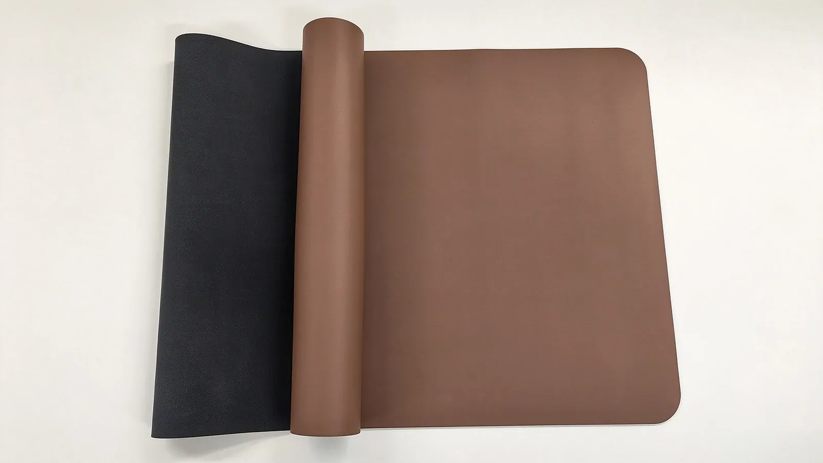 A coffee-colored yoga mat with one end rolled up and the rest lying open on a pale table, with a dark mat at the left
