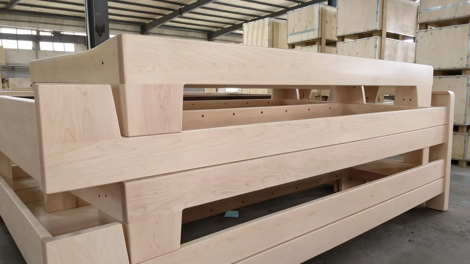 Several pale light-toned wooden Pilates bed frames stacked in the workshop, with wooden packing crates behind them