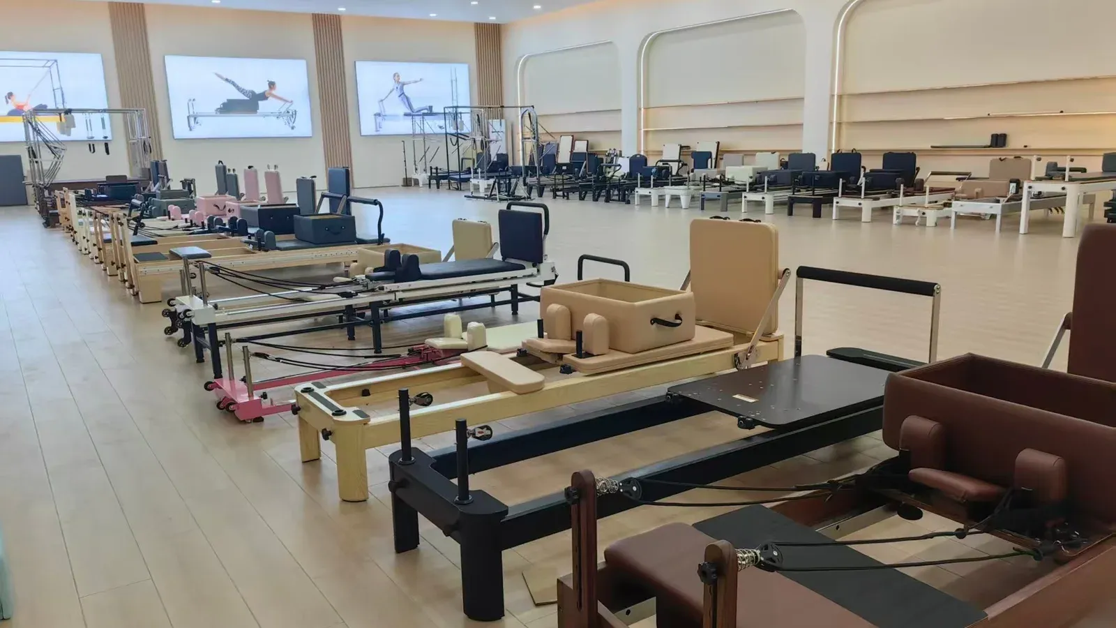 Pilates reformer showroom floor with rows of reformers in different frame and leather finishes, wall screens behind and a trapeze frame on the left