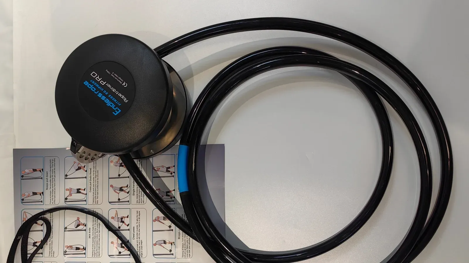 A black magnetic resistance unit with a coiled black coated rope, a blue sleeve, carabiners and an exercise poster laid beside it
