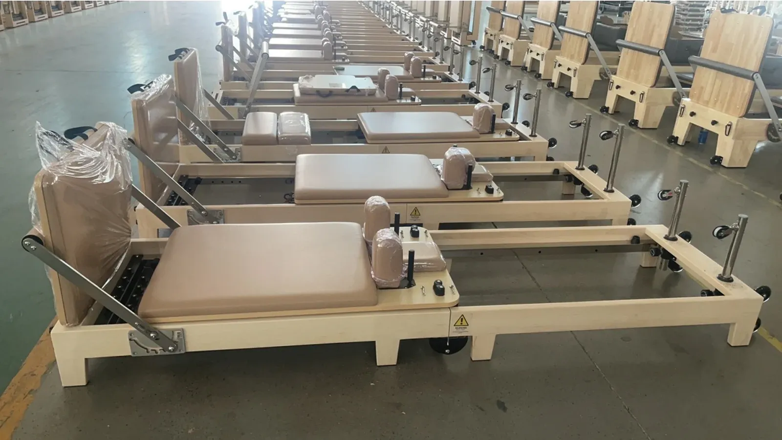Rows of maple foldable Pilates reformer units on a factory floor, with pale wood frames, tan upholstered pads and castors under the base