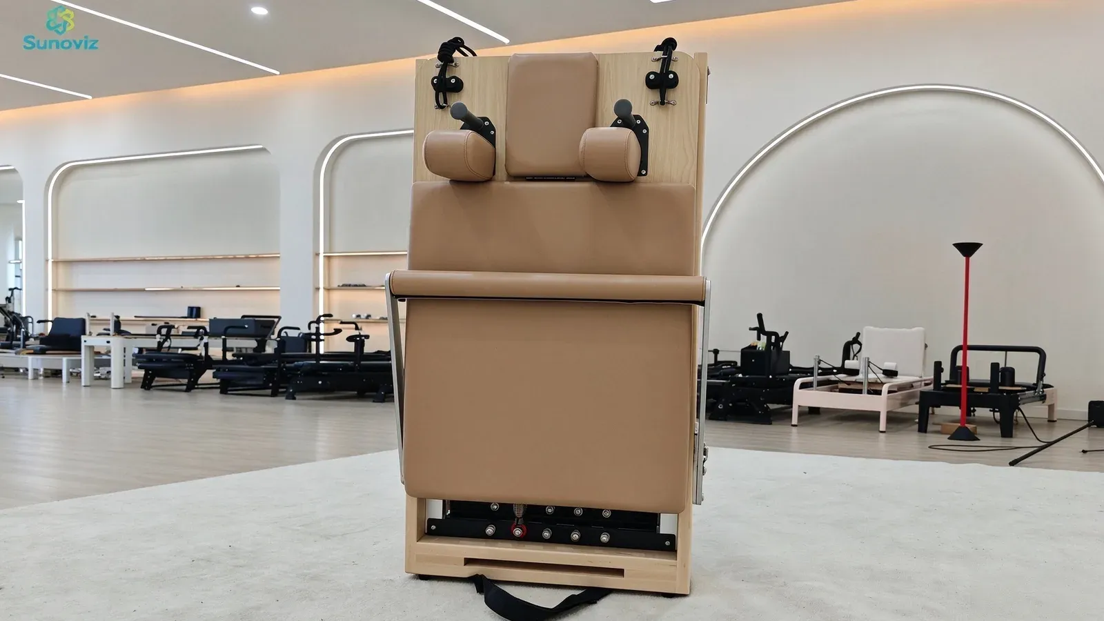 A foldable maple Pilates reformer standing upright in its folded storage position on castors, with a carry handle below and other equipment behind in a showroom
