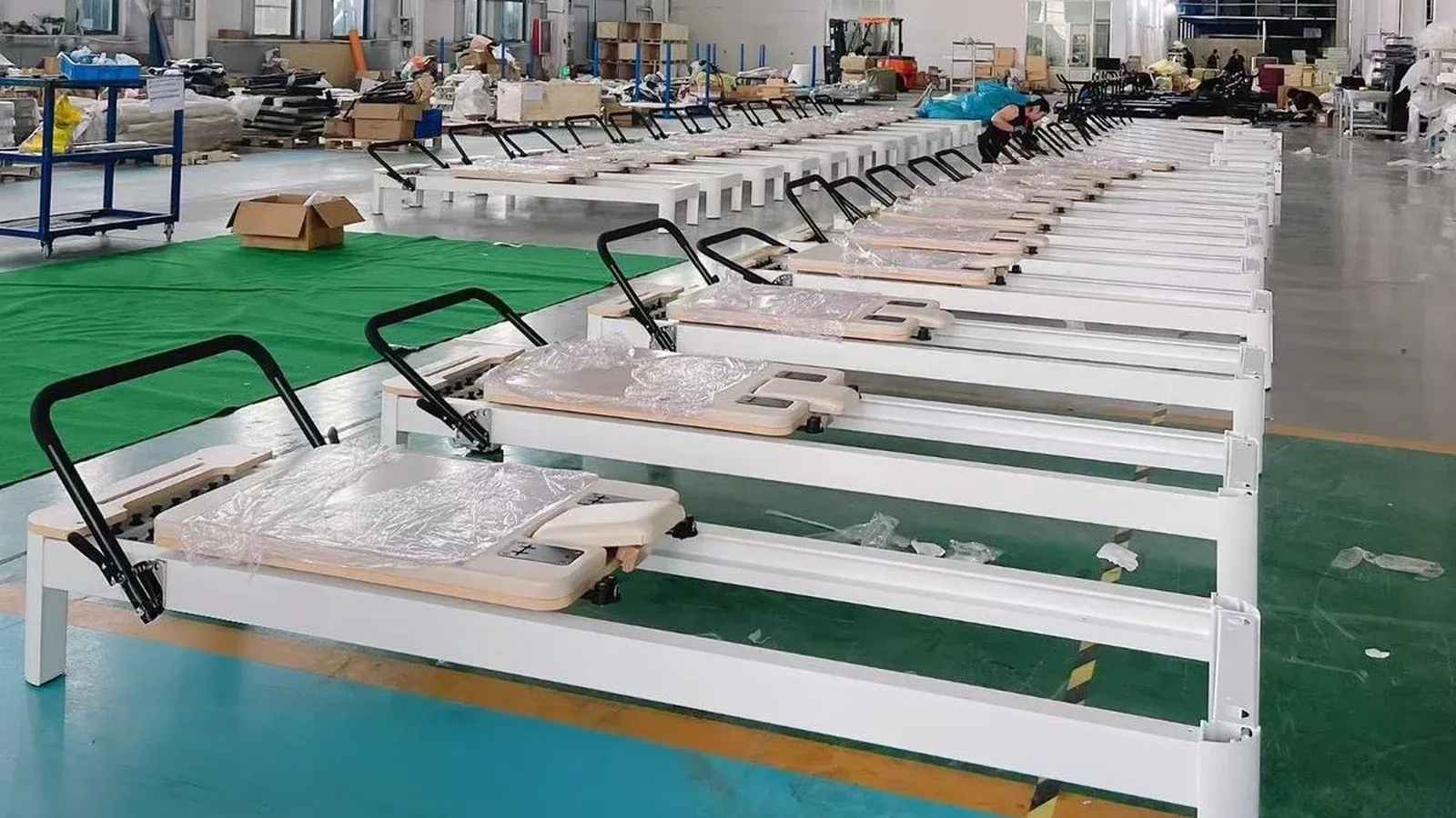 A long row of white-framed Pilates reformers in the workshop, each with a black curved handle at the near end and a cream-beige upholstered carriage wrapped in clear protective film