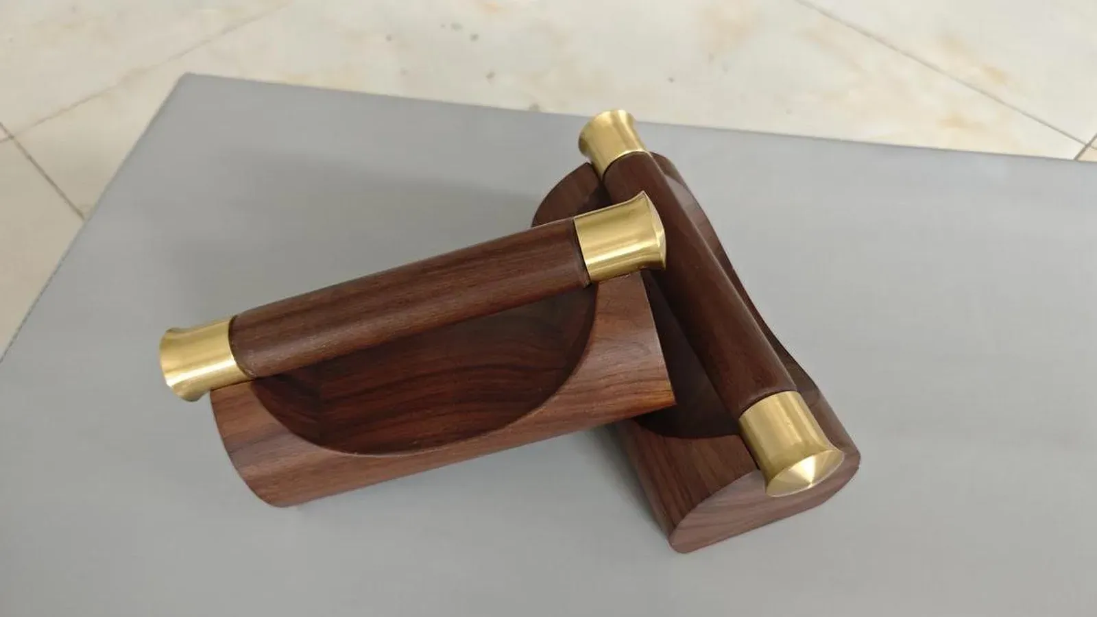 Two solid walnut push-up stands with copper-coloured caps on the handle ends resting crossed on a light grey surface, one showing the curved scooped wooden base