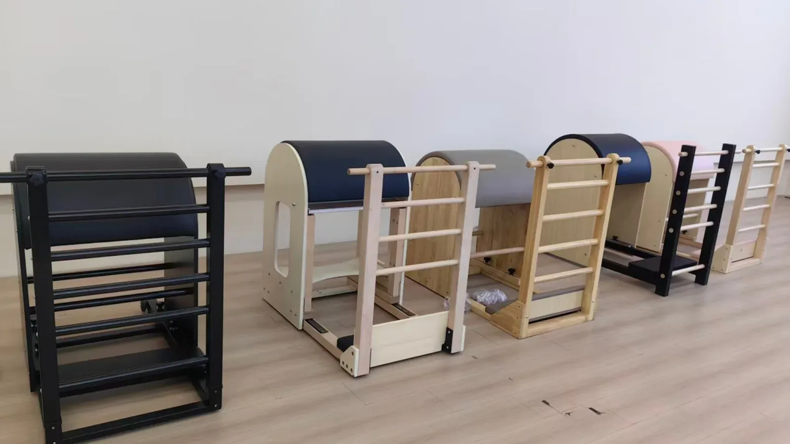 Row of five wooden Pilates ladder barrels on a light wood floor, with padded barrels in black, navy, grey and pink over timber and black ladder frames