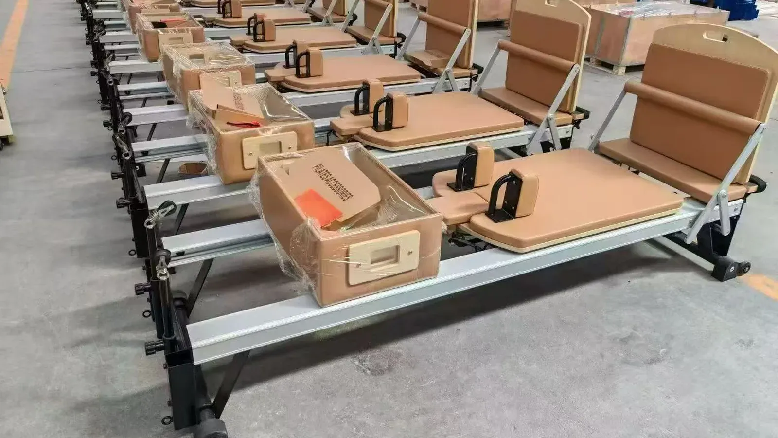 A row of tan-upholstered Pilates reformers on a workshop floor, each carriage carrying a film-wrapped box, with packing crates and a truck behind