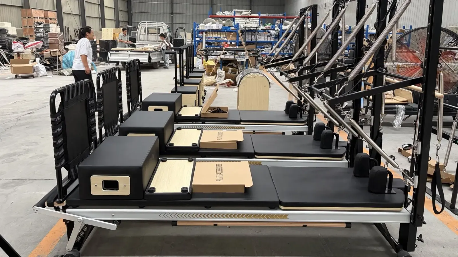 Row of Pilates reformers on a factory floor, each fitted with a tall half tower of springs and straps at the near end, with black upholstery on the carriages