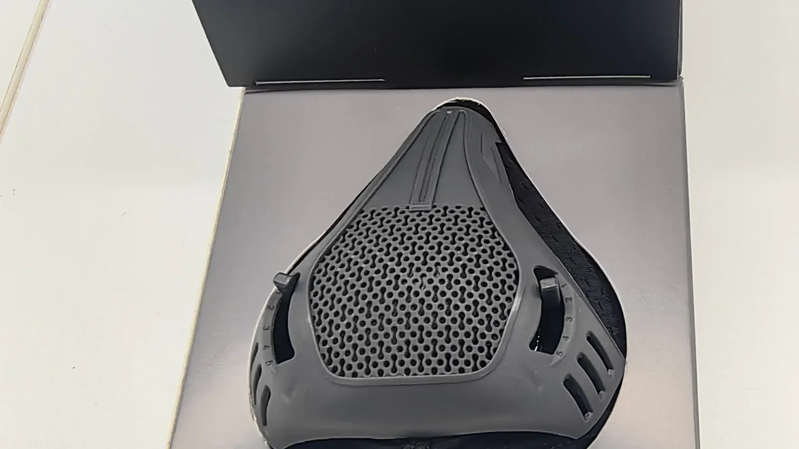 A black resistance sports training mask in its open presentation box, showing the mesh front panel and an adjustable side vent