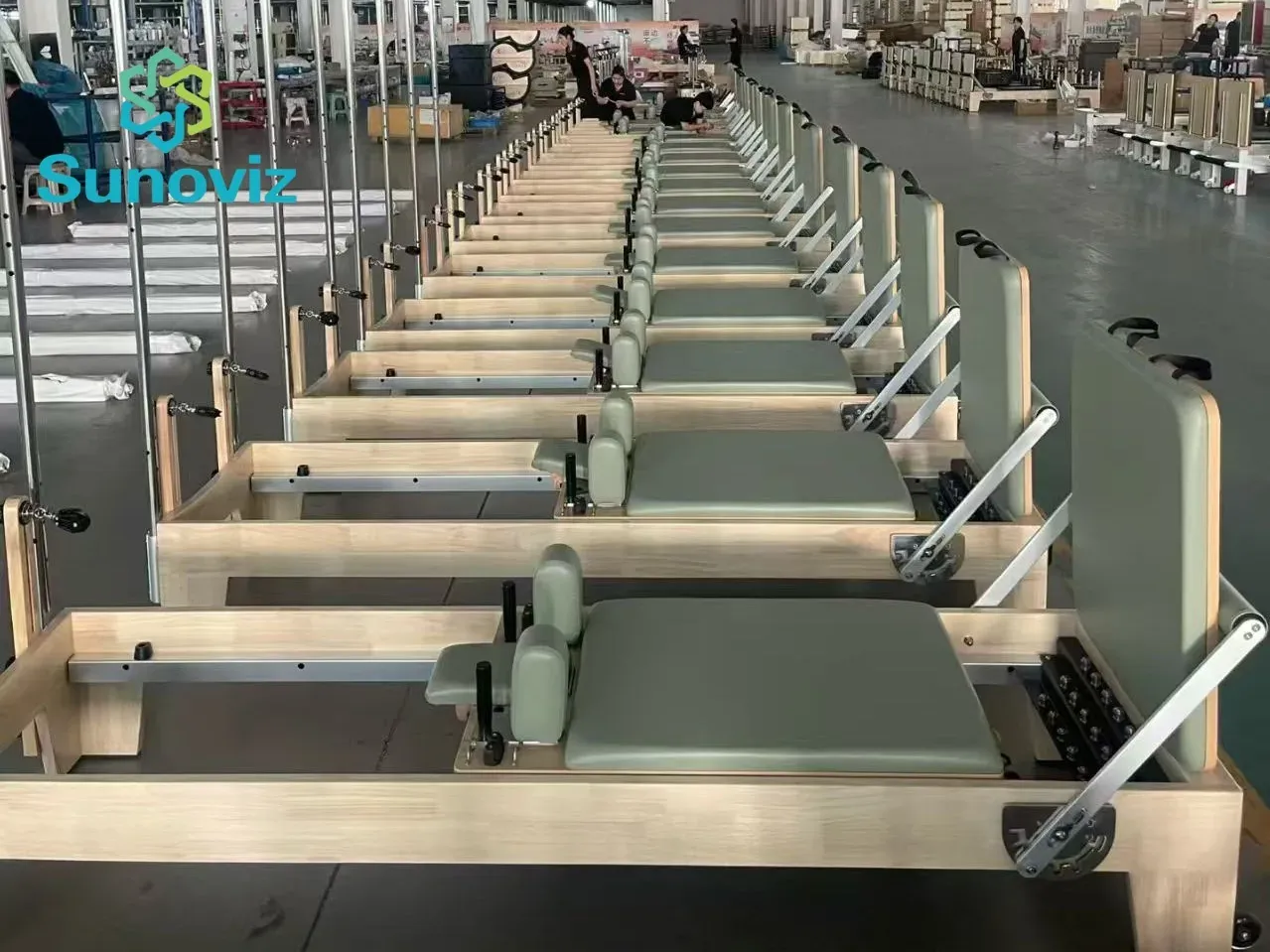 Row of wooden Pilates reformers with pale maple frames and sage green leather upholstery in the factory