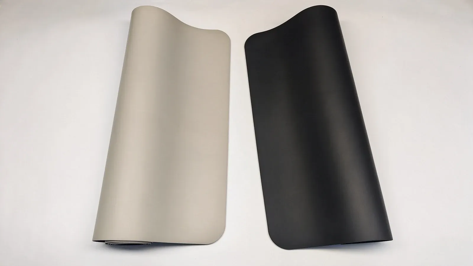 A beige yoga mat and a black PU rubber yoga mat side by side on a pale surface, the beige one with one end curled at the lower left