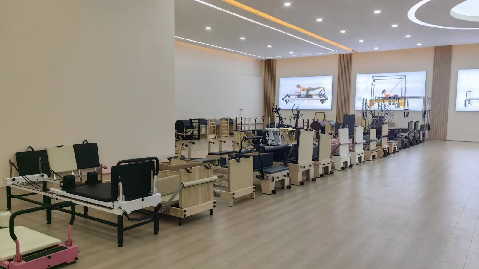 Pilates showroom with a pink-framed unit in the foreground and a row of equipment along the left, including a reformer with a dark carriage on a black-and-white frame and a pale timber chair