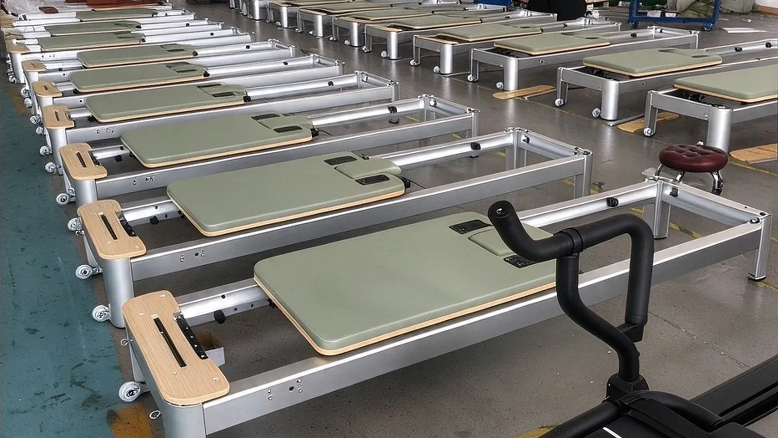 Rows of sage green classic Pilates reformers on a factory floor, with silver metal frames, timber end blocks and castors