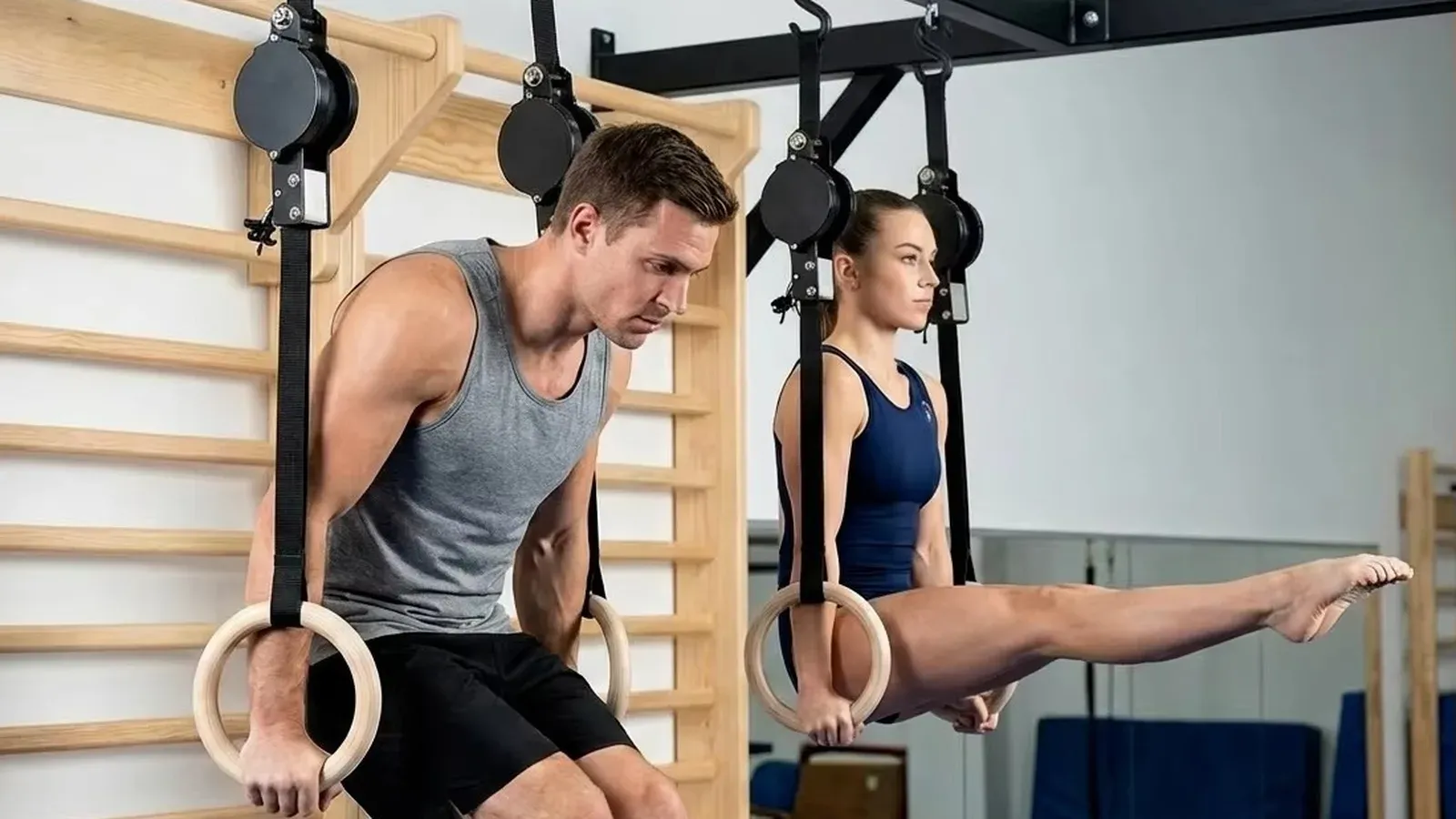 Two athletes training on wooden gymnastic rings hung from black straps with circular adjustment housings, beside a wooden wall ladder