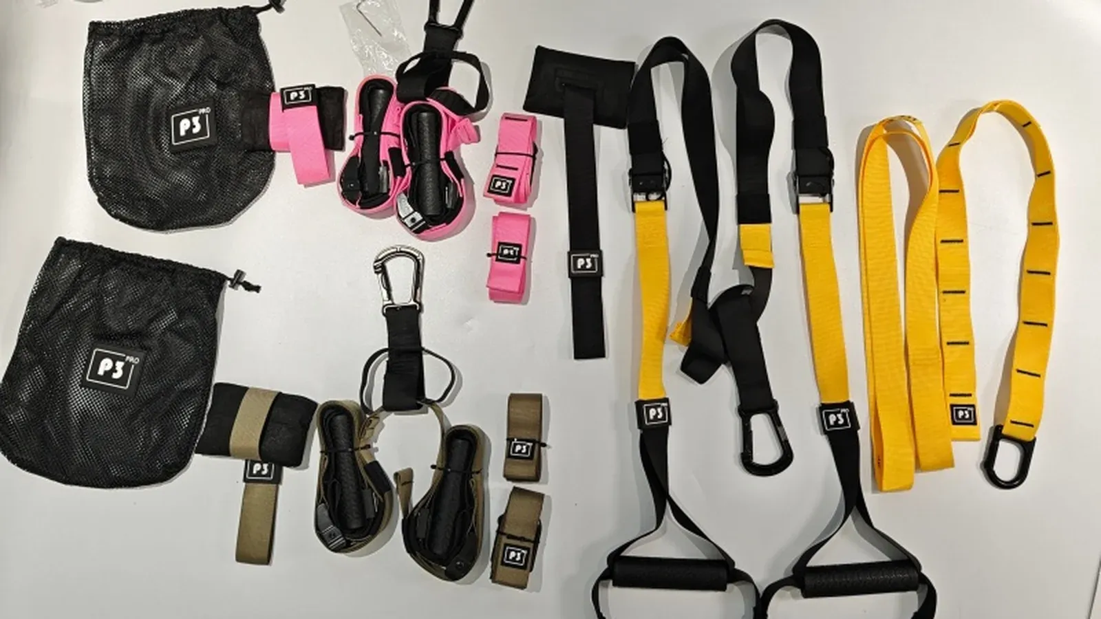 Suspension trainer sets laid out in army green, pink and yellow, with metal carabiners, padded handles and black mesh carry pouches
