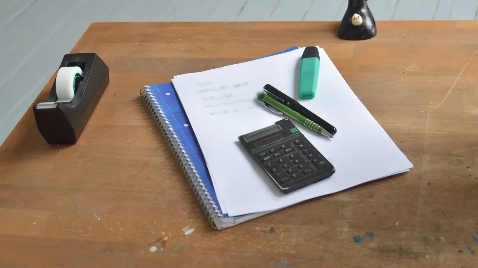 Handwritten notes on a notebook with a calculator, a highlighter and pens on a wooden desk
