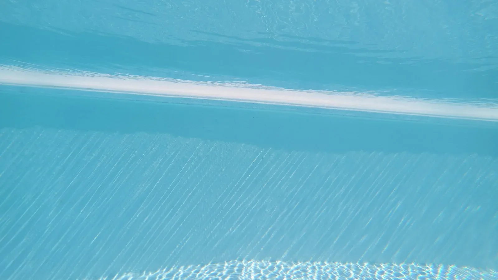 Underwater view up a pool wall toward the waterline with sunlight from above