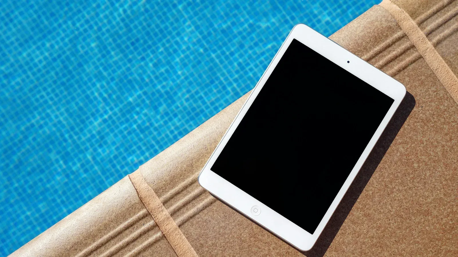 Tablet with a dark screen lying on stone coping at the edge of a pool