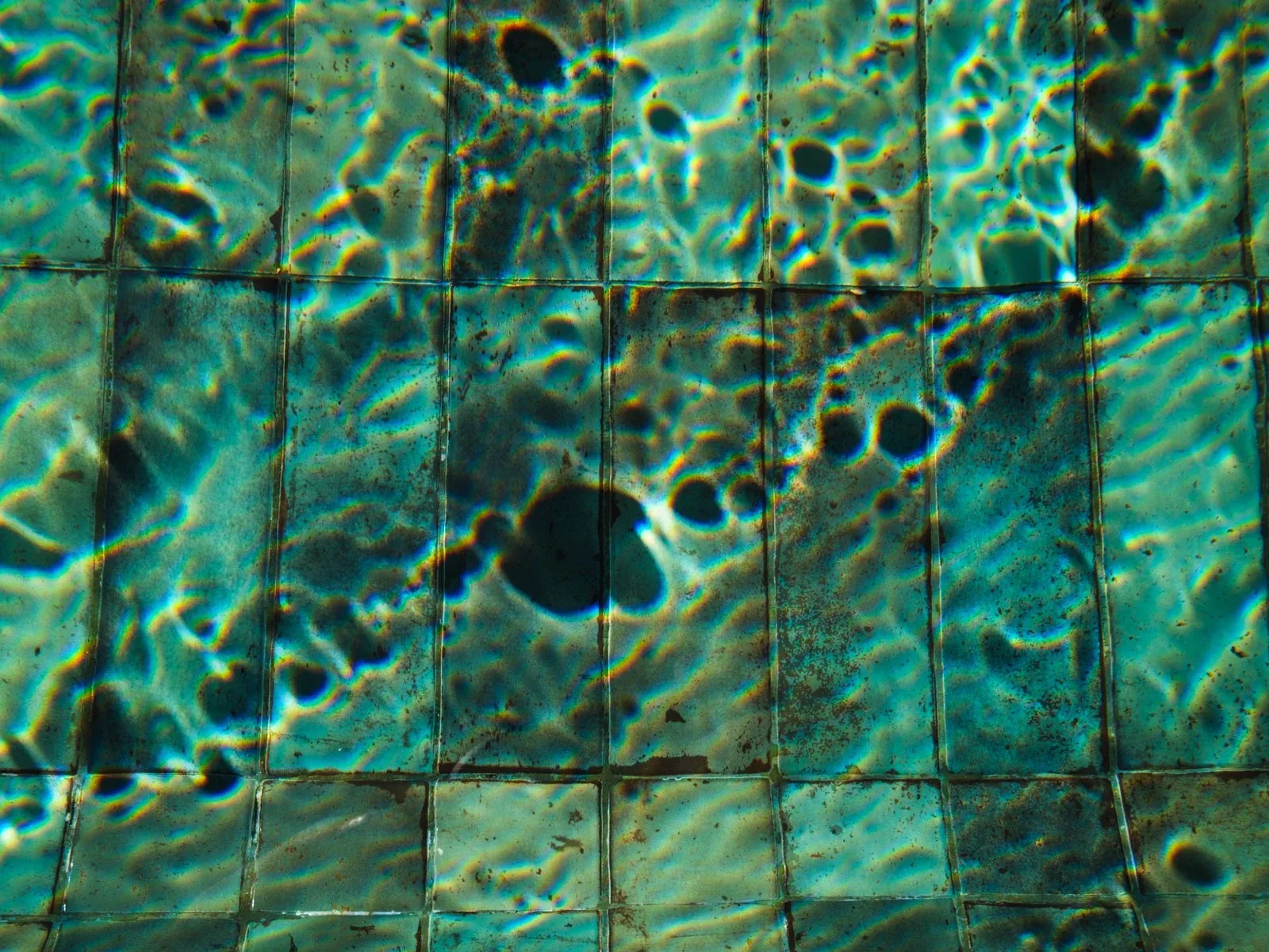 Green-tinted pool tiles under rippling water with dark spots of algae