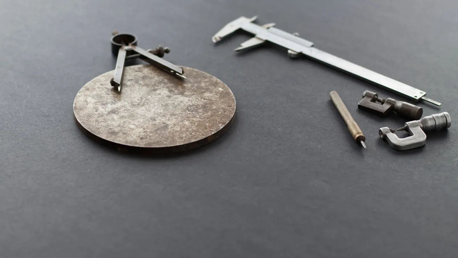 Vernier caliper, micrometers and a compass beside a machined metal disc on a grey surface