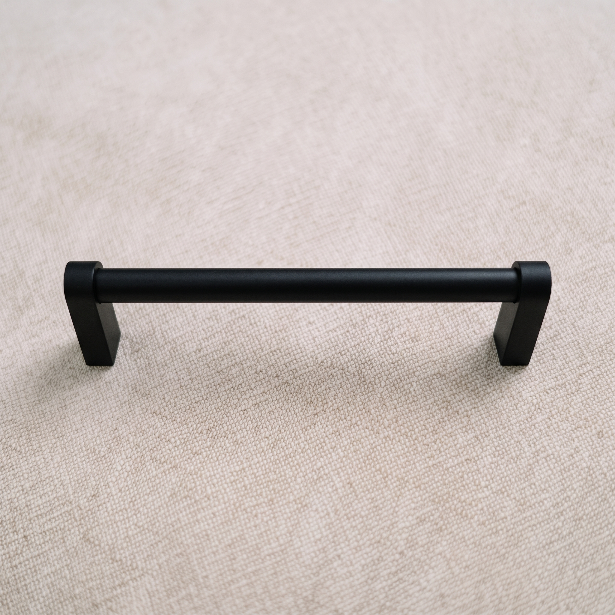 Front elevation of a matt black smooth cylindrical aluminum bar pull on pale linen, with square block feet below the horizontal bar