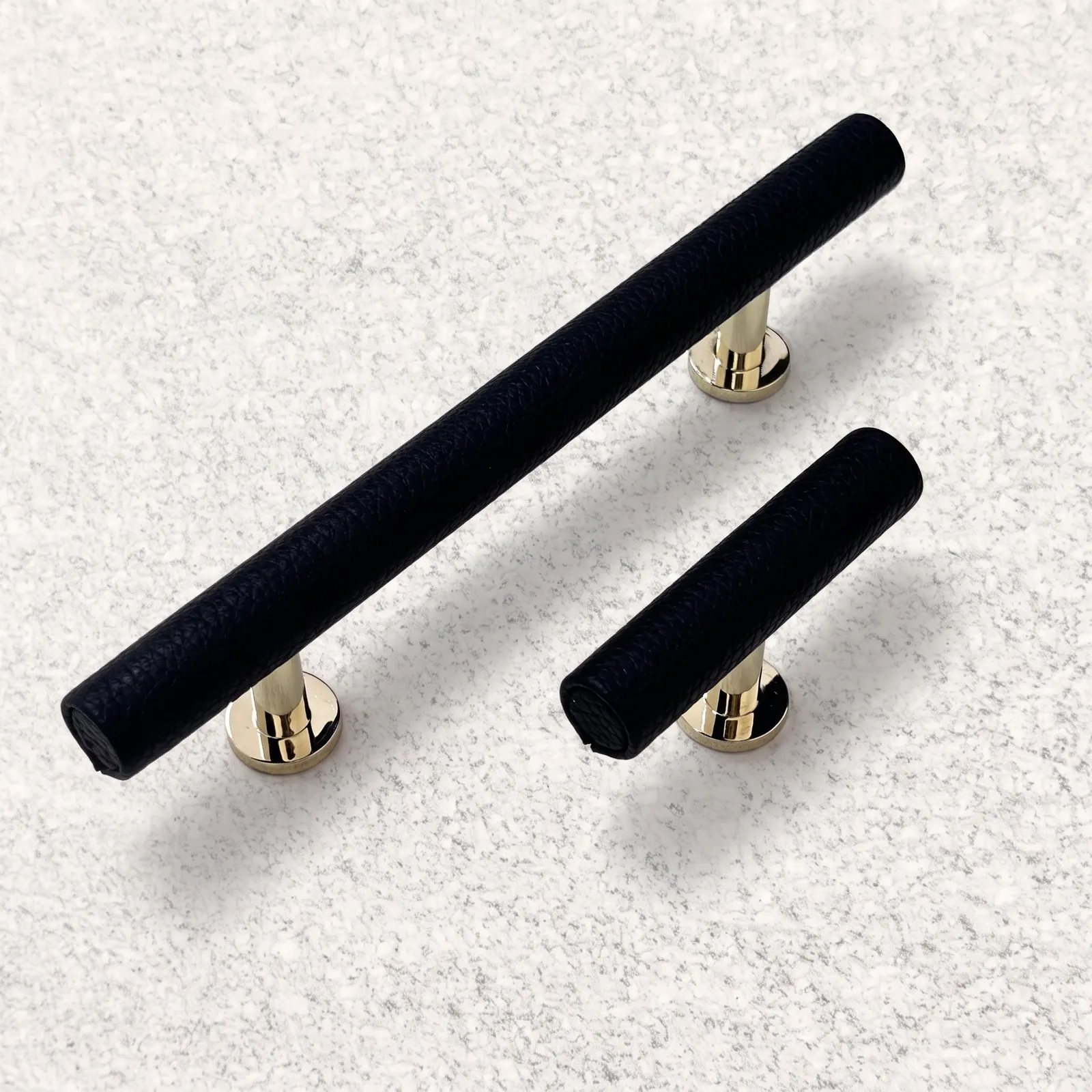 Angled view of black leather-wrapped T-bar pulls with polished gold-toned posts on a pale textured surface.