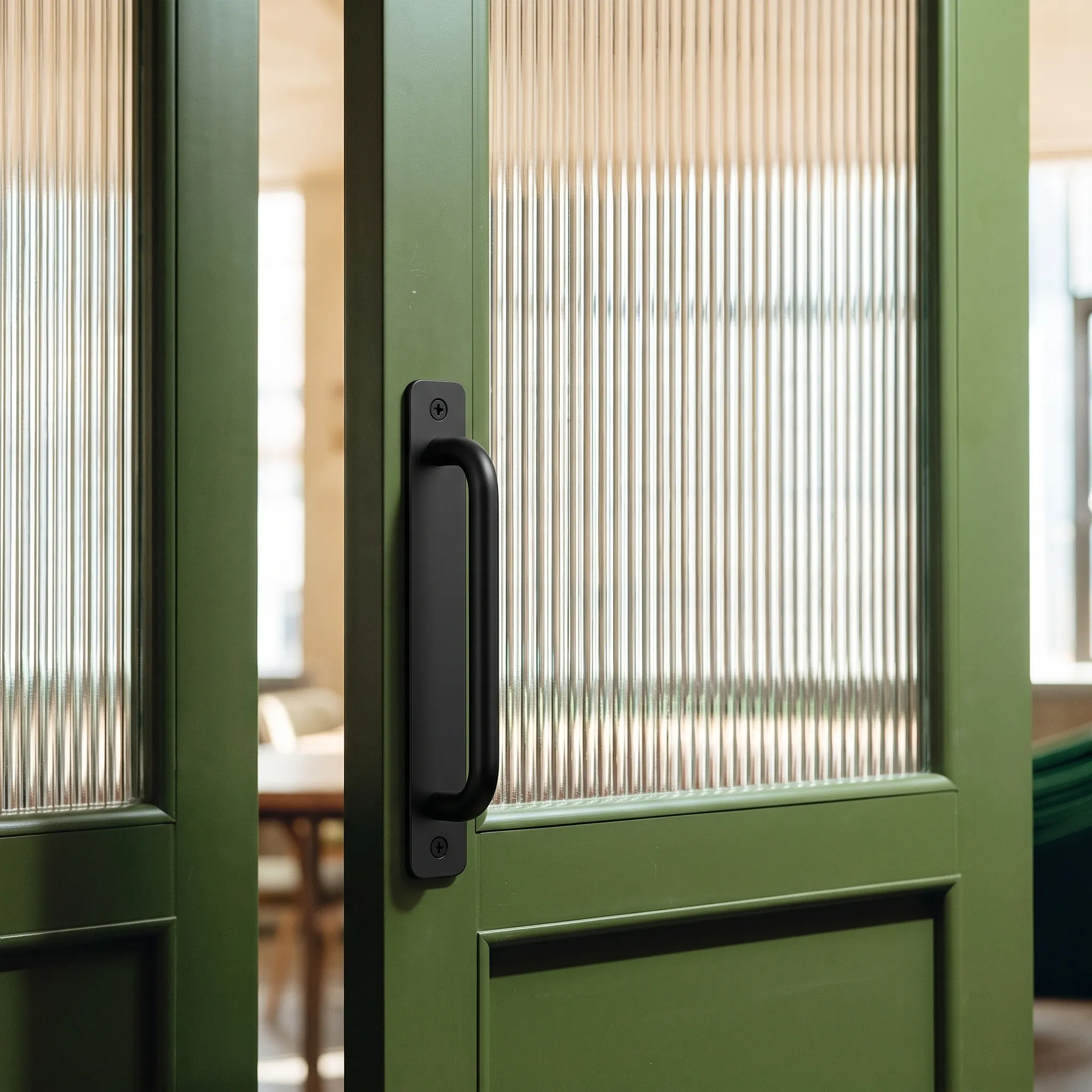 Matt black U-bar plate pull on a green door with a reeded glass panel, its mounting screws visible on the flat plate while the door stands ajar.