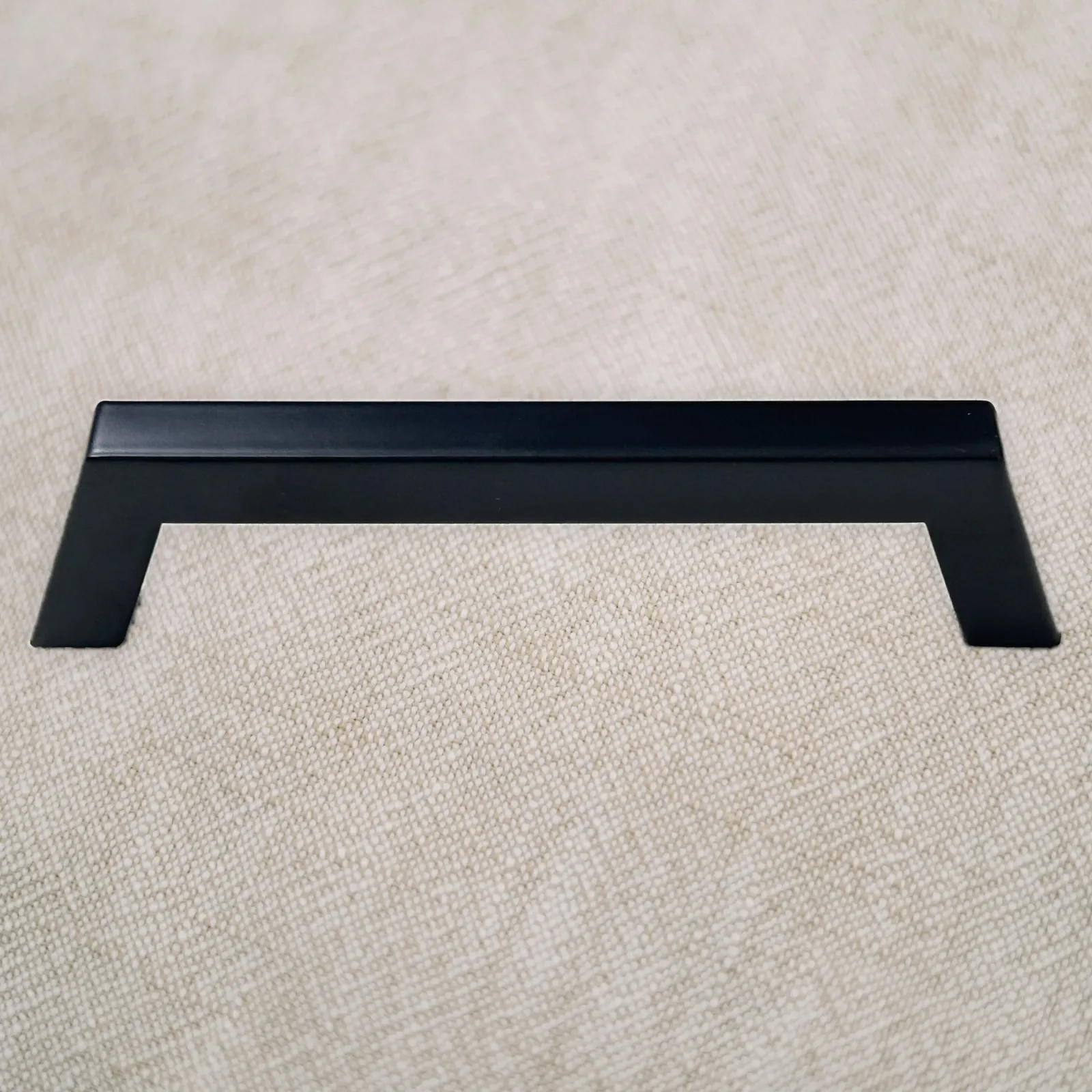 Front view of the Matt Black trapezoidal stainless steel cabinet pull standing on natural linen