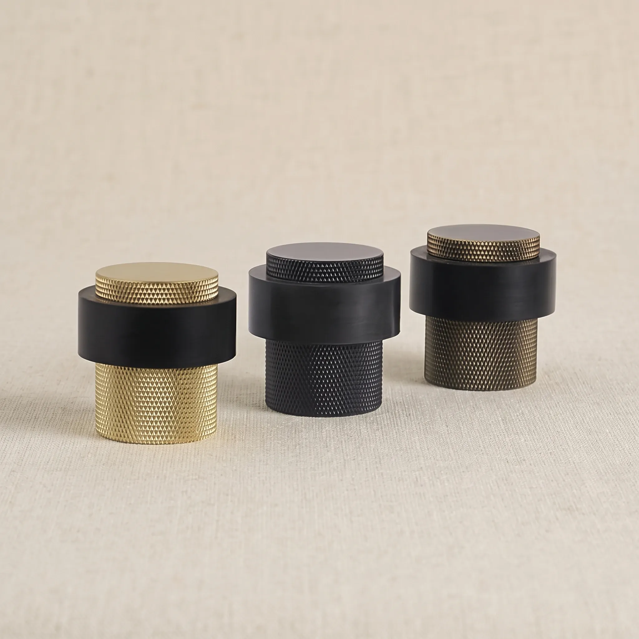Lineup of the knurled cylindrical door stop in Brass Gold, Matt Black, and Antique Bronze finishes on a warm neutral studio surface.
