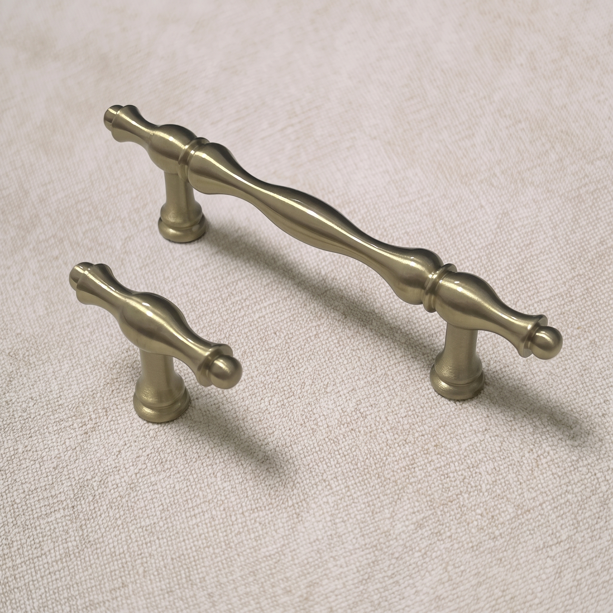Angled view of a wavy brass bar pull with hourglass mounting feet next to its matching T-knob on a pale linen surface.