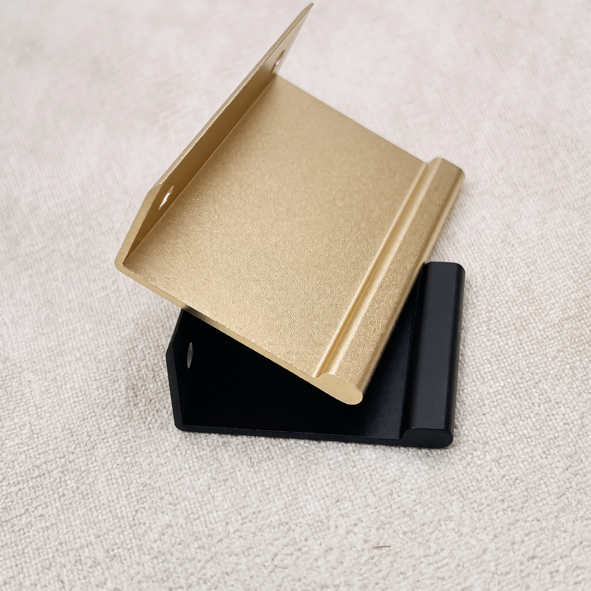 Close-up of a champagne gold smooth-arc edge pull resting across a matt black piece on a pale beige fabric surface, showing the rounded nose along the folded edge.
