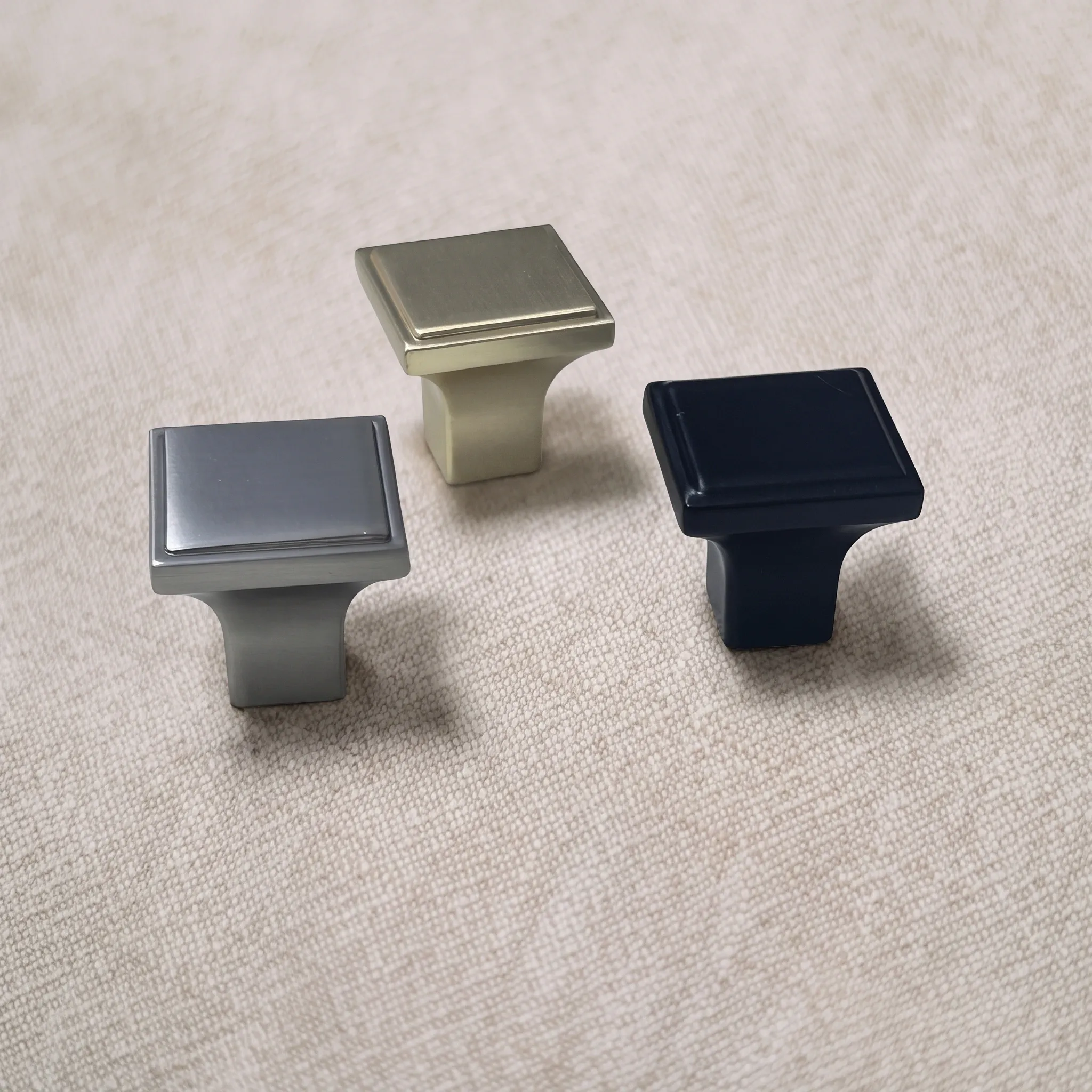 Three upright square cabinet knobs in Satin Nickel, Brush Gold, and Matt Black with stepped faces on a textured beige studio surface.