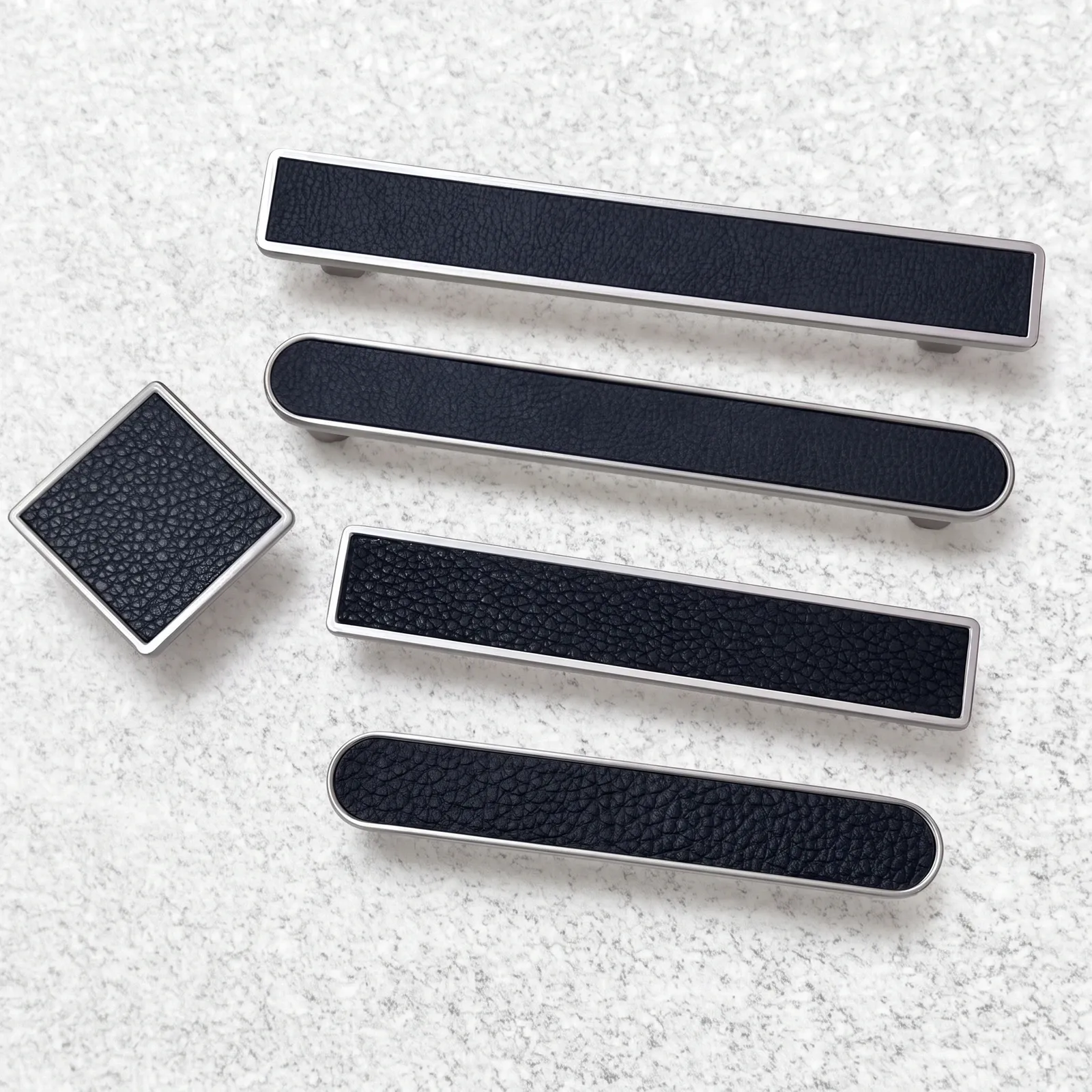 Overhead view of framed black leather bar pulls in polished silver-toned borders with a matching square leather knob on a pale textured surface.