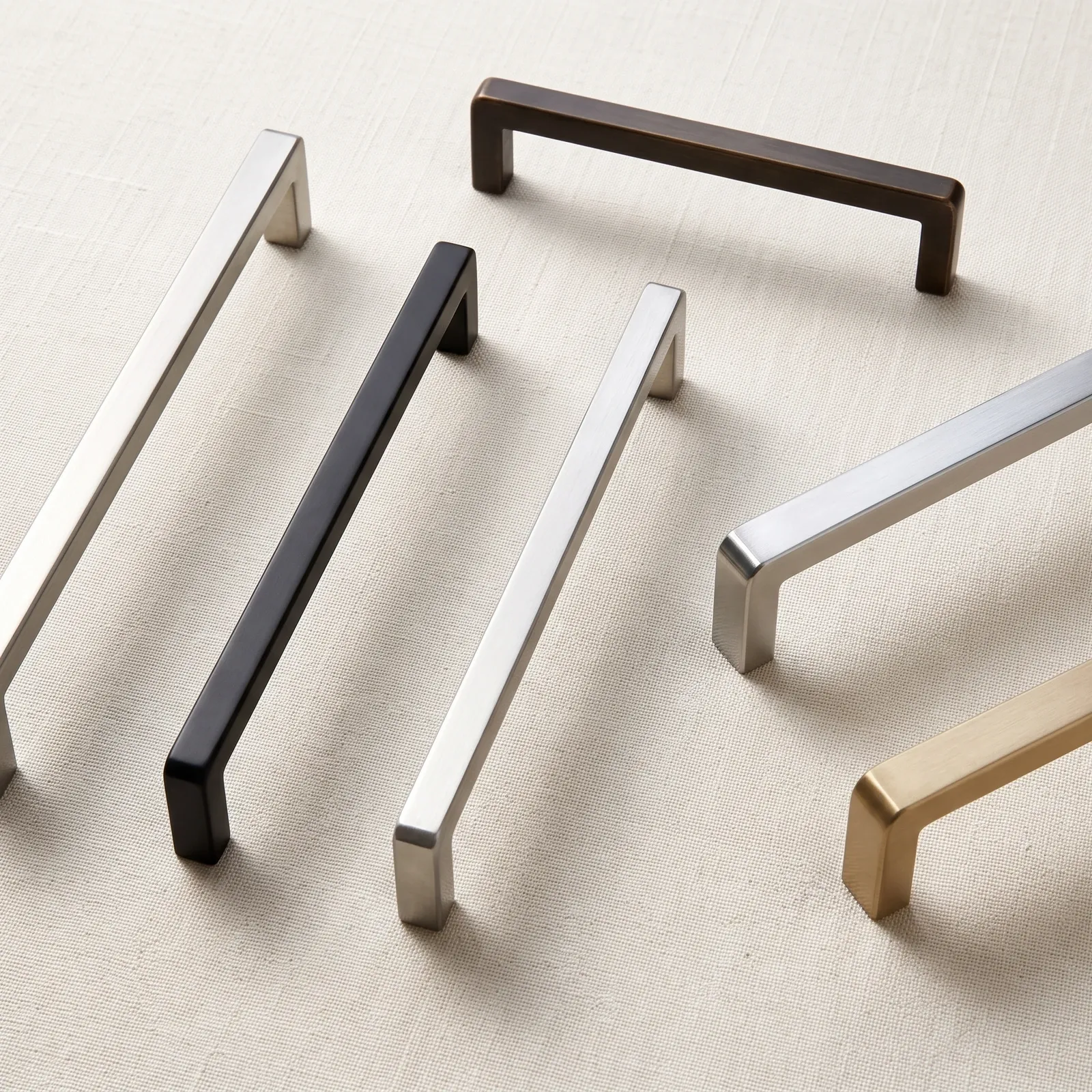 Square bar cabinet pulls in a range of metallic finishes arranged on linen