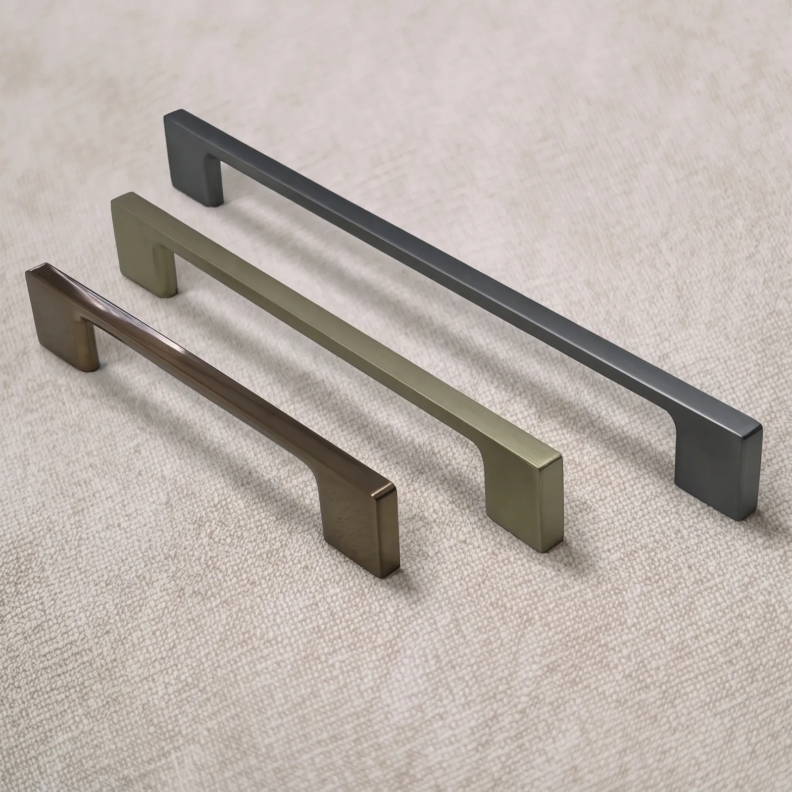 Three D-profile zinc alloy bar pulls in dark, gold, and bronze tones with straight bars and flared feet on natural linen