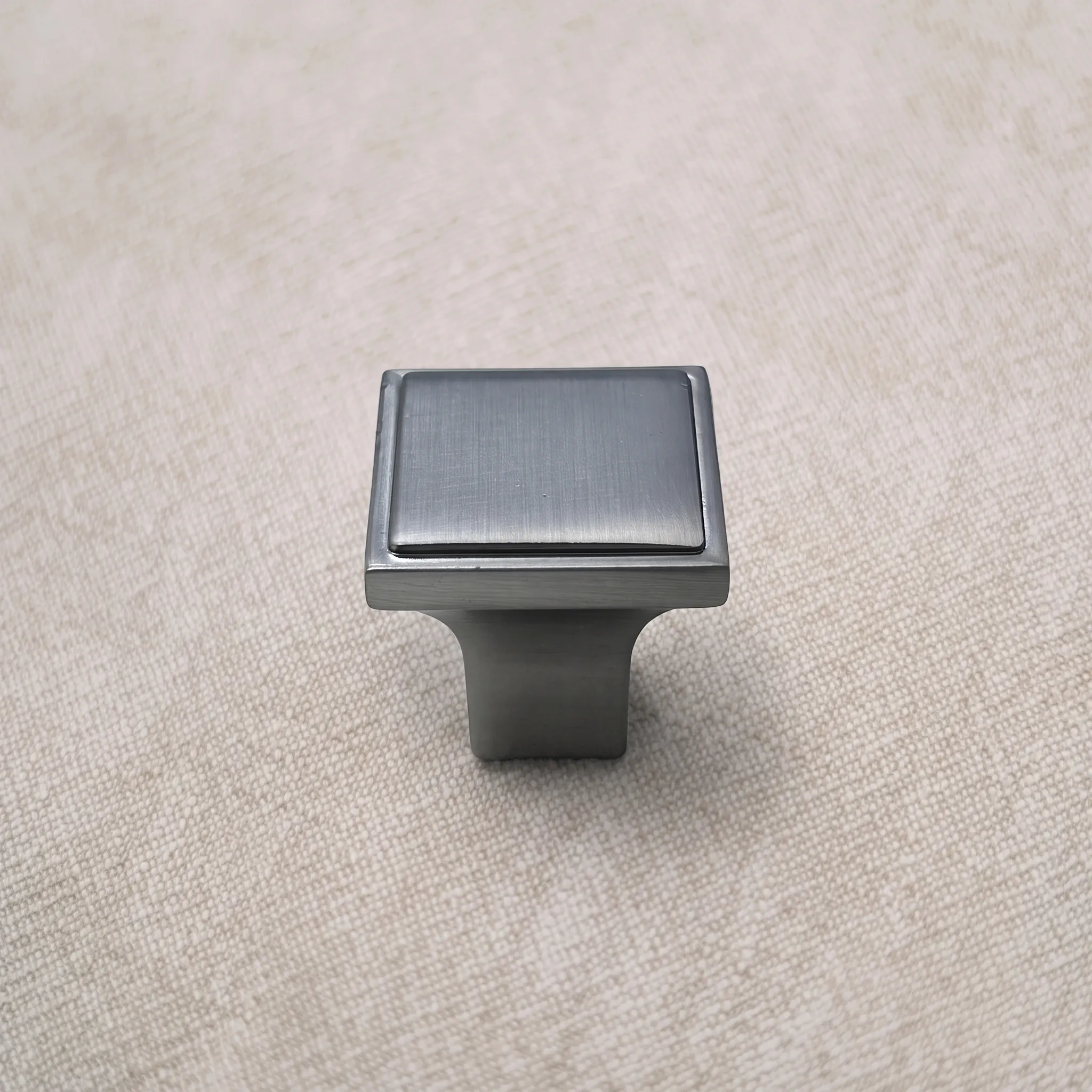 Angled studio shot of a Satin Nickel square cabinet knob highlighting the raised inner panel and narrow pedestal on textured beige surface.