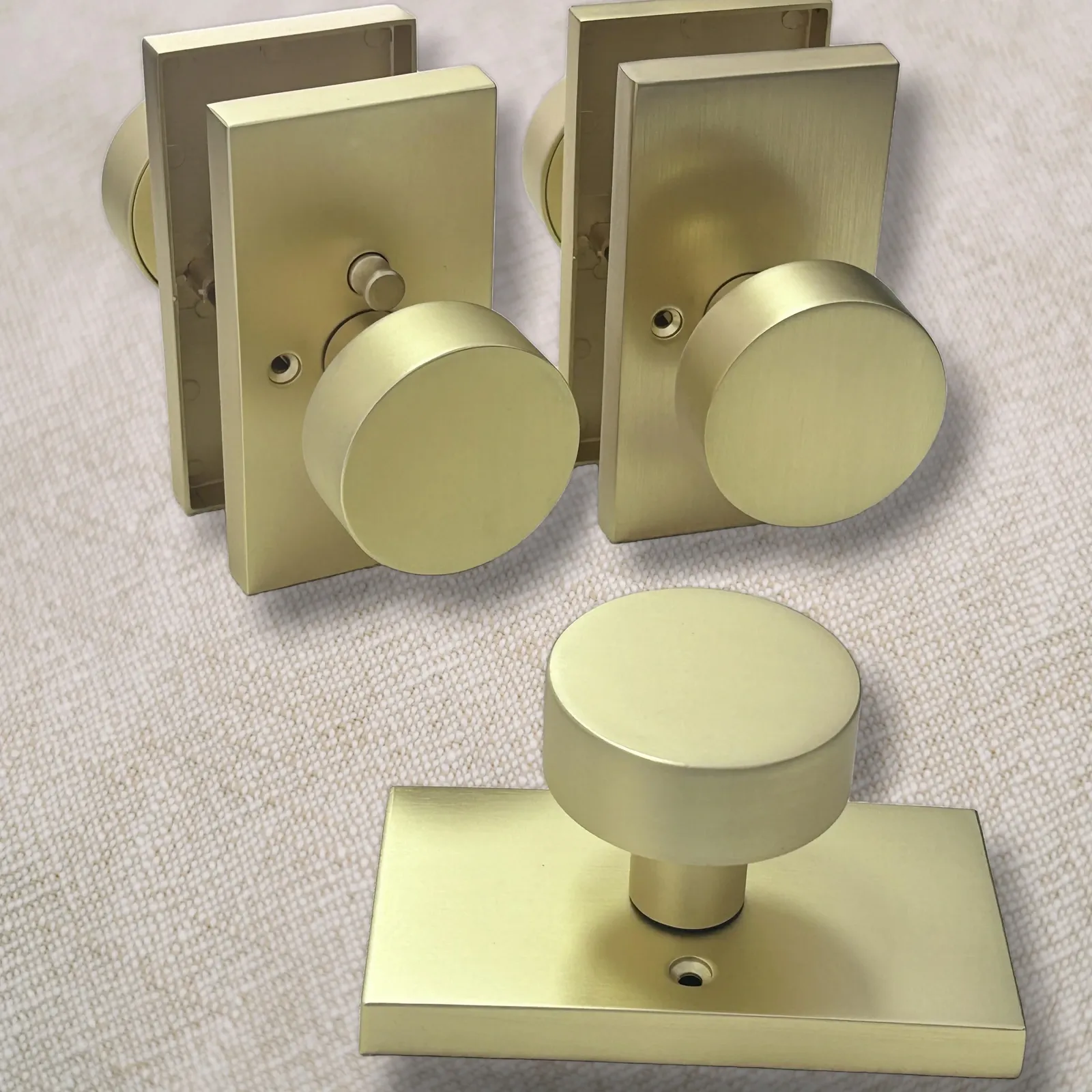 Three brushed gold door knob assemblies with rectangular rose plates on linen