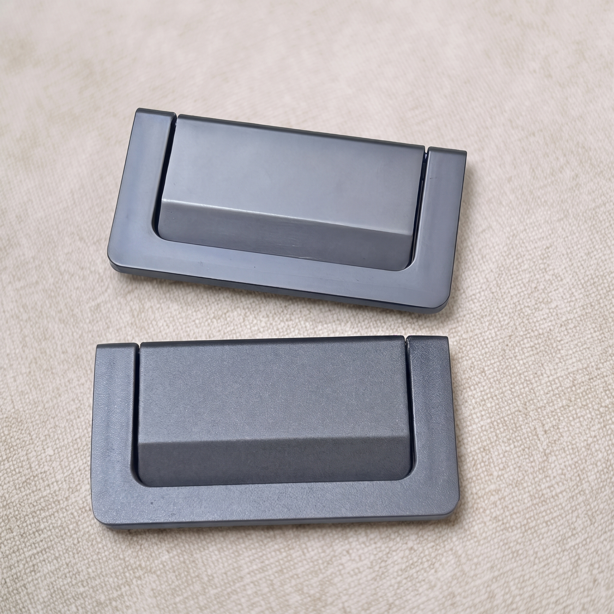 Two graphite grey recessed flip pulls staggered on a pale beige textured surface, each with the finger tab resting flush inside the plate.