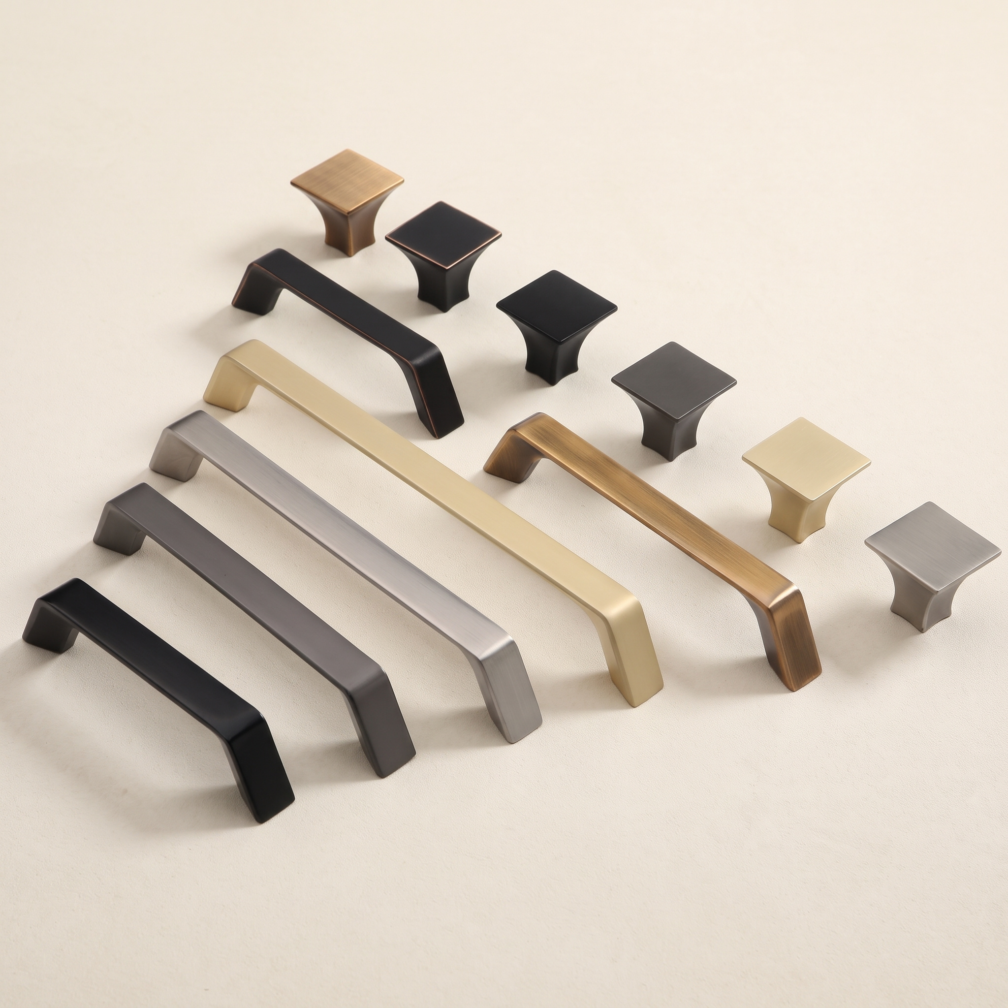 Rows of chamfered-end zinc alloy cabinet pulls arranged over a plaster wall in matt black, copper-toned, brushed gold, brushed nickel and grey finishes