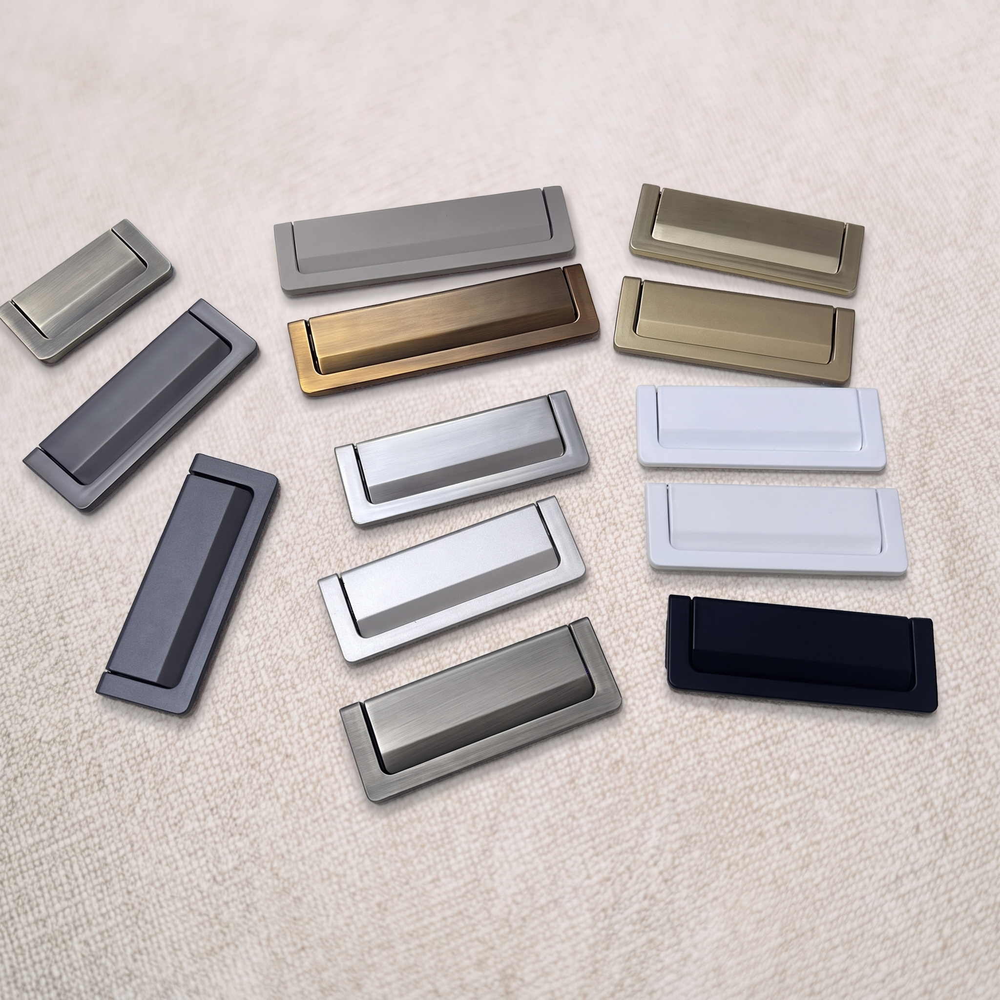 Recessed flip pull laid out across its full finish range, with dark, bronze, brass, satin and white units fanned on a pale beige textured surface.