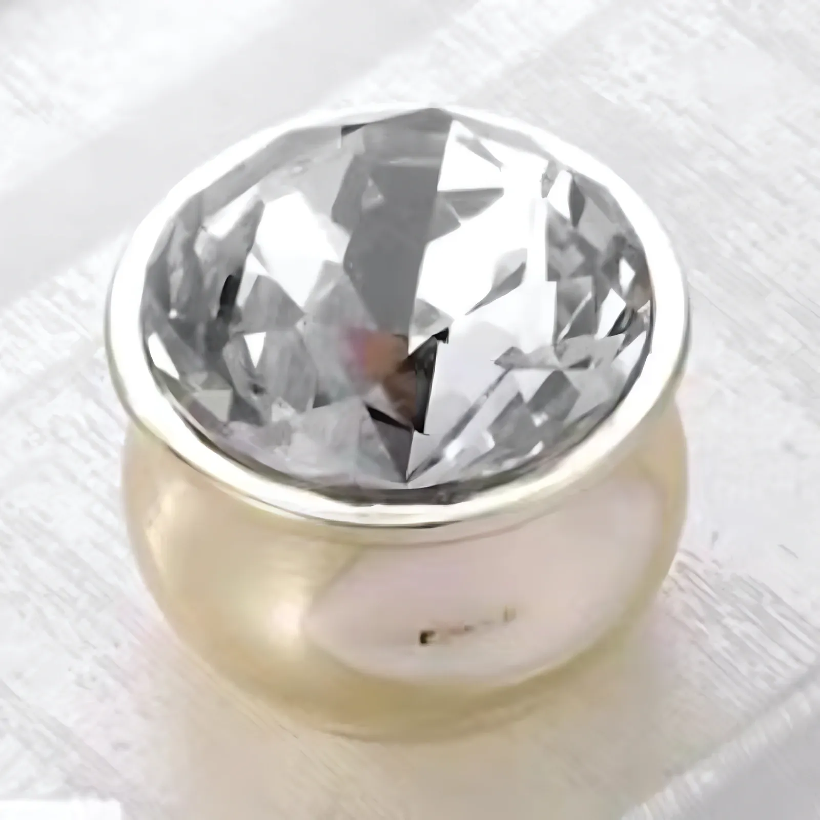 Angled view of one round brilliant-cut clear crystal knob set in a smooth gold-tone cup-shaped base, standing on white painted wood.