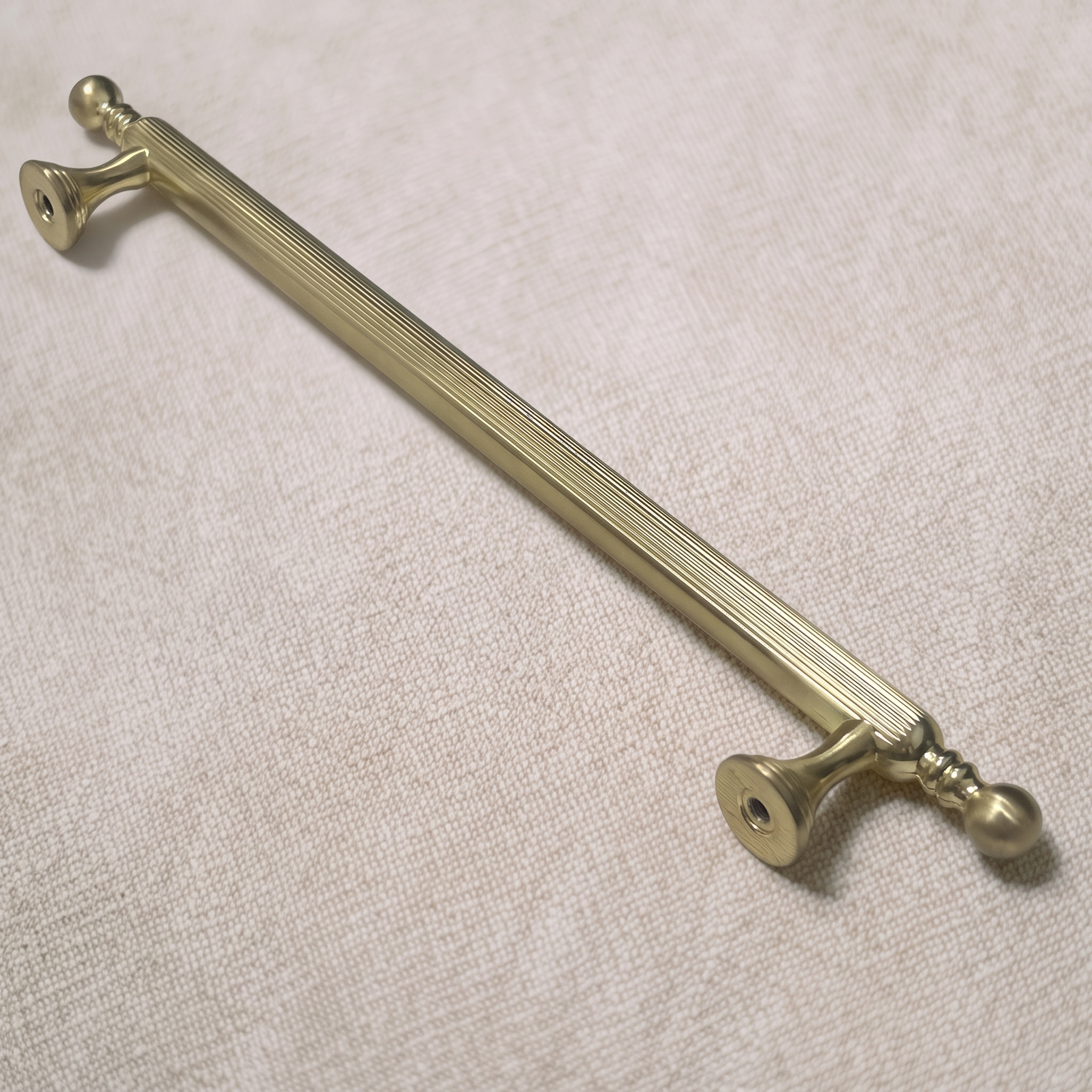 Three-quarter view of the straight-grain zinc cabinet pull in Brass Gold across a pale linen surface, showing the full bar and both bell feet