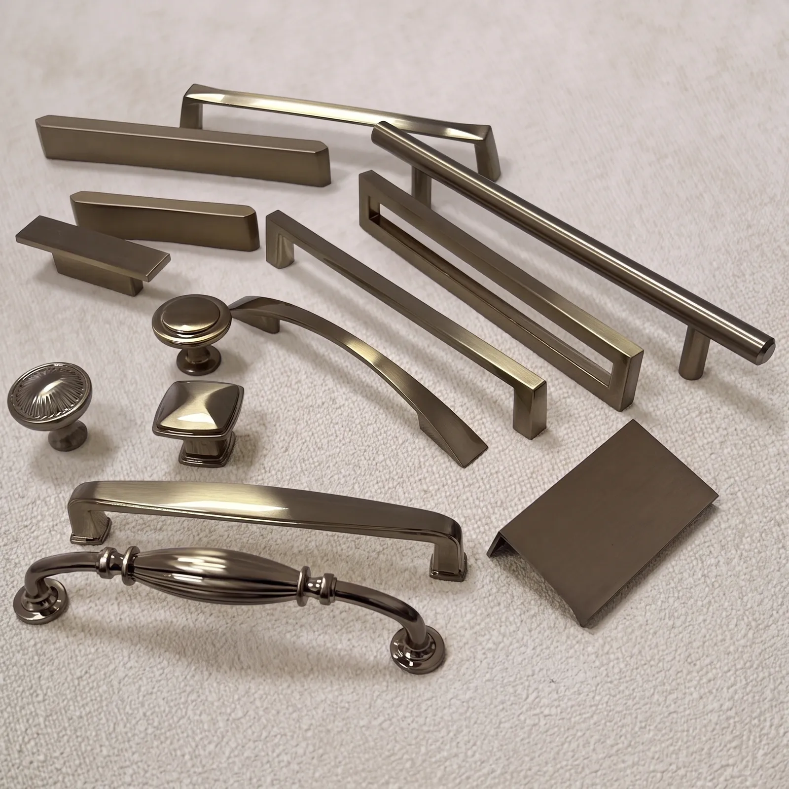 Overhead view of an aromatherapy gold furniture hardware group on a cream towel: bar pulls, an open frame pull, an arch pull, a slim T-bar, a bent-plate edge pull, round knobs, a square knob and a pull with a fluted centre band