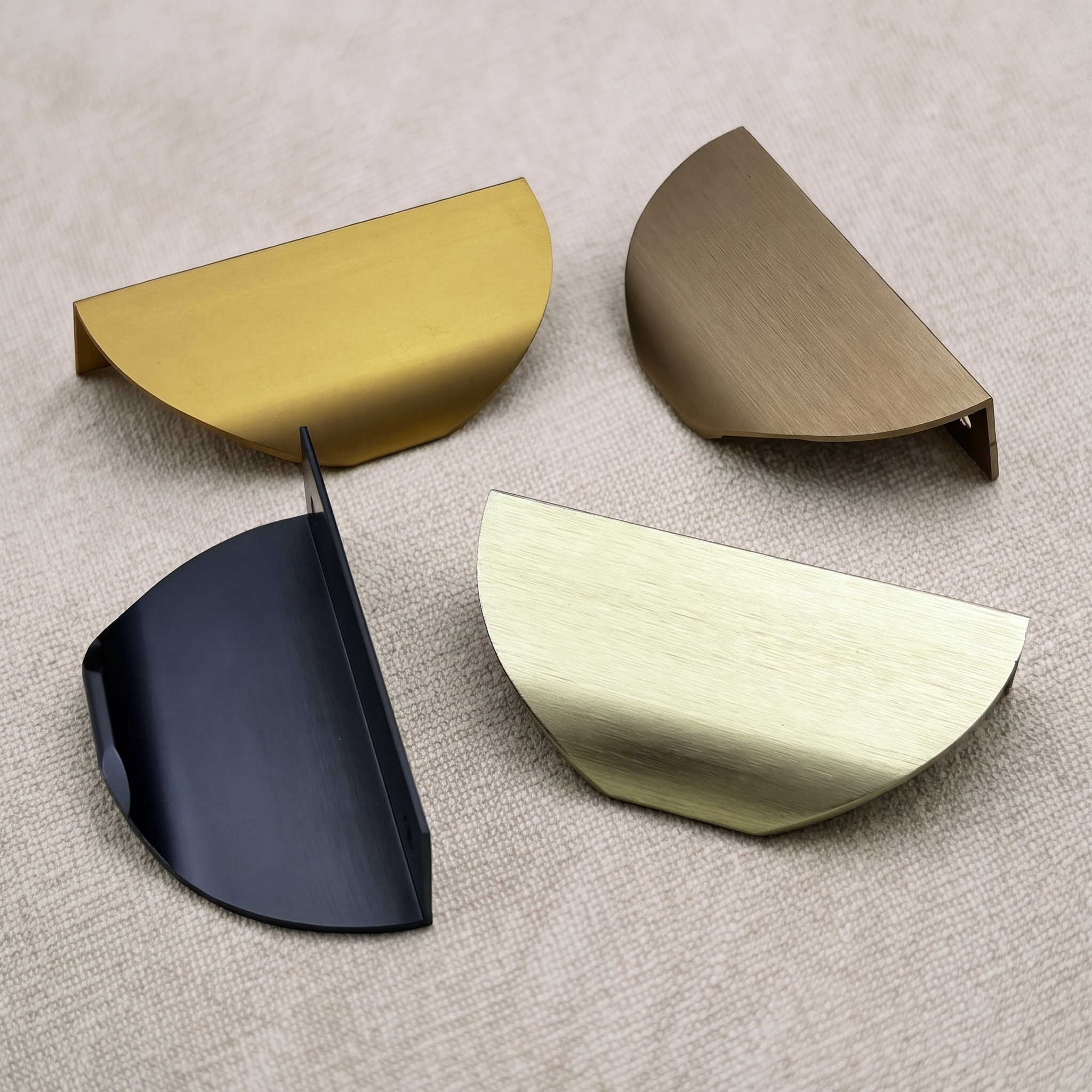 Four half-moon edge pulls from the range laid flat on a pale linen surface, showing the Brush Gold, Matt Black, Brass Gold and Champagne finishes